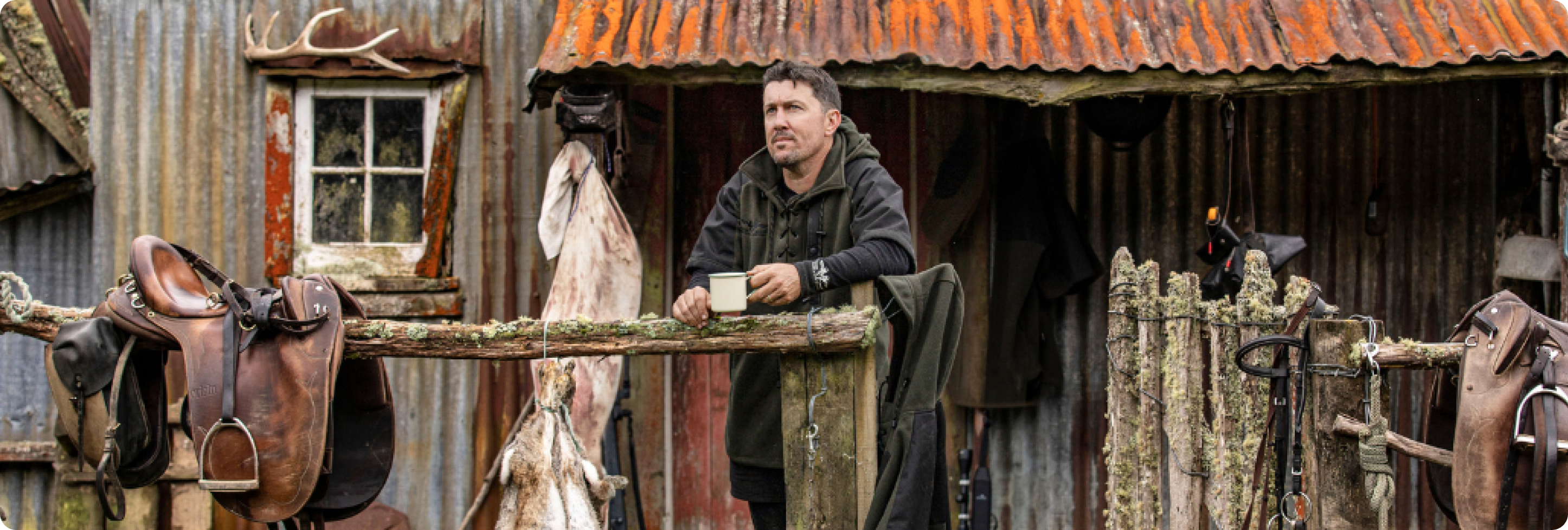 A man wearing Game Gear attire drinks a coffee outside a hunting hut in New Zealand.