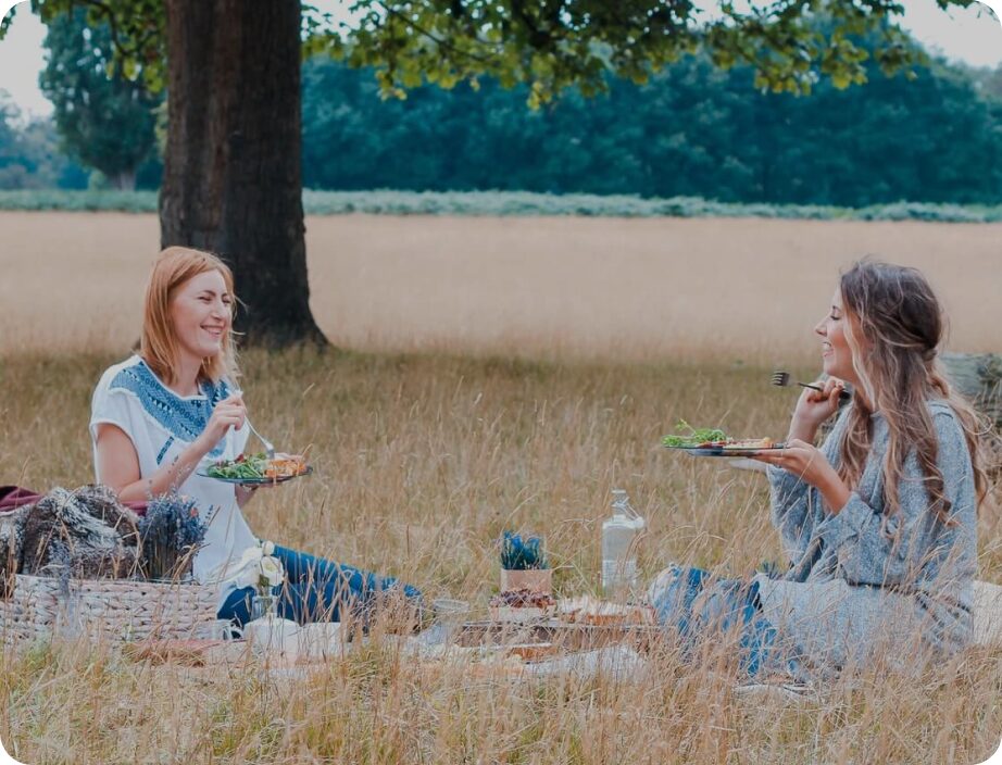 Two women enjoy a picnic in a field with a tree in the background.